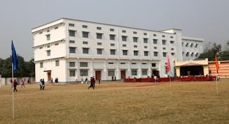 A large multi-story white building with numerous windows situated on a spacious grassy field. A group of people, including some wearing uniforms, are walking and gathering around. The building features a small stage area set up with red chairs and several colored flags on poles positioned across the field.