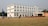 A large multi-story white building with numerous windows situated on a spacious grassy field. A group of people, including some wearing uniforms, are walking and gathering around. The building features a small stage area set up with red chairs and several colored flags on poles positioned across the field.