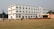 A large multi-story white building with numerous windows situated on a spacious grassy field. A group of people, including some wearing uniforms, are walking and gathering around. The building features a small stage area set up with red chairs and several colored flags on poles positioned across the field.