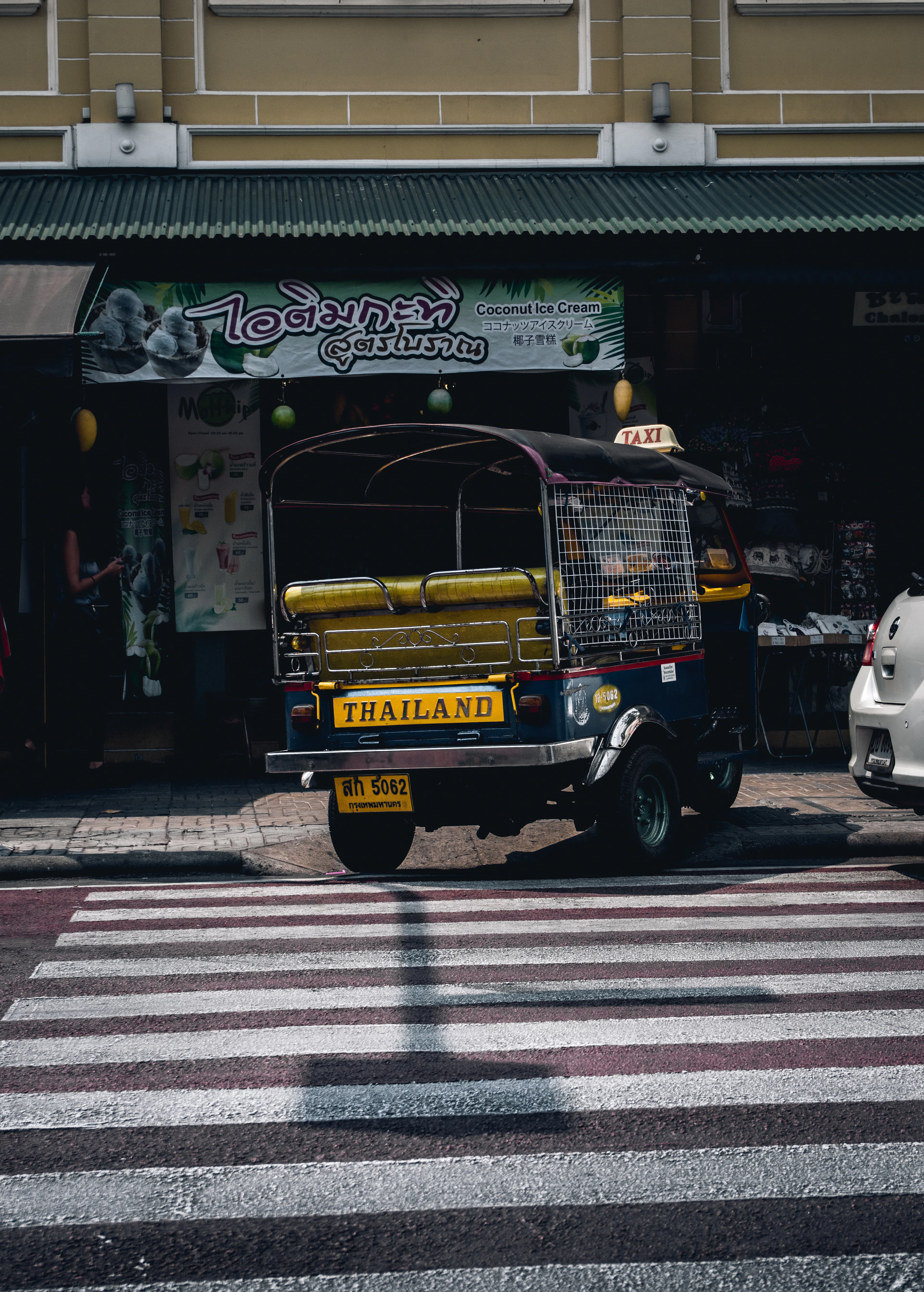 A tuk-tuk marked 'Thailand' is parked at a crosswalk outside a Bangkok shopfront.