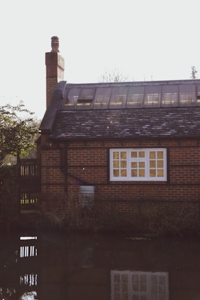 A brick house with a sloped roof featuring skylights and a prominent chimney sits by a tranquil water body. The house has a large window with white framing and a garden area to the side. There is some greenery and reflections of the house in the water, creating a serene atmosphere.