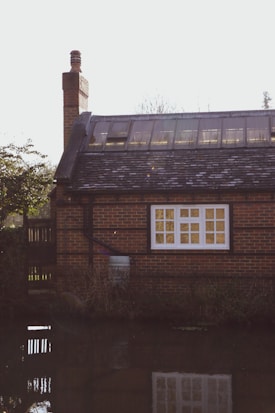 A brick house with a sloped roof featuring skylights and a prominent chimney sits by a tranquil water body. The house has a large window with white framing and a garden area to the side. There is some greenery and reflections of the house in the water, creating a serene atmosphere.