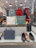 A display of handbags and shoes in a retail store. There are three handbags on a shelf: one blue, one red, and one white, all with different designs. Below them, a blue handbag is displayed alongside three pairs of flat shoes in black, pink, and blue. The background shows a retail store setting with a person in the background.