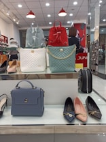 A display of handbags and shoes in a retail store. There are three handbags on a shelf: one blue, one red, and one white, all with different designs. Below them, a blue handbag is displayed alongside three pairs of flat shoes in black, pink, and blue. The background shows a retail store setting with a person in the background.