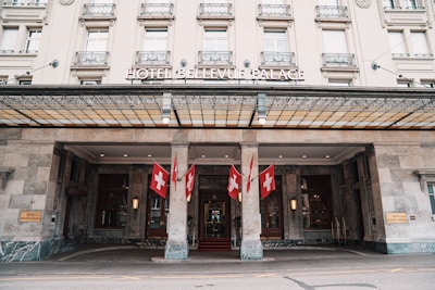 A grand hotel entrance features a classical facade with ornate architectural details. Multiple Swiss flags are prominently displayed, enhancing the building's elegance. The entrance canopy is supported by stone columns, and there are wall-mounted lights near the doors.
