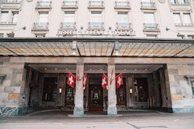 A grand hotel entrance features a classical facade with ornate architectural details. Multiple Swiss flags are prominently displayed, enhancing the building's elegance. The entrance canopy is supported by stone columns, and there are wall-mounted lights near the doors.