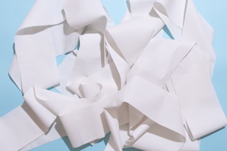 A tangled heap of white paper towel or tissue strips set against a light blue background. The paper appears textured and creates a complex pattern with folds and layers.
