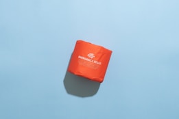 A bright orange package labeled 'Emergency Roll' is placed on a light blue background. The package has bold white text and subtle shadows cast underneath.