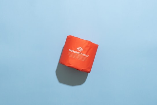 A bright orange package labeled 'Emergency Roll' is placed on a light blue background. The package has bold white text and subtle shadows cast underneath.