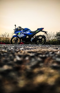 A blue motorcycle parked on a road, with an overcast sky in the background and some foliage visible behind it. The motorcycle features prominent logos and has a sporty design.