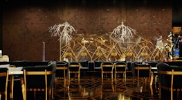 A luxurious and sophisticated interior space featuring elegant dining tables and chairs with black upholstery and wooden accents. The walls have a reflective, geometric design with intricate, illuminated floral decorations. The floor showcases a polished, patterned surface, contributing to the high-end atmosphere.