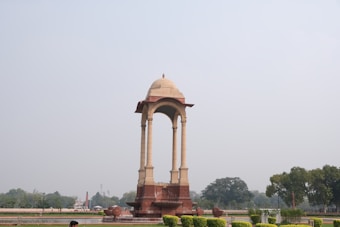 An ornate architectural canopy structure stands prominently in an open landscaped area. The structure is supported by four tall pillars and topped with a dome. Surrounding the canopy are well-maintained lawns and trees visible in the background, under a clear sky.