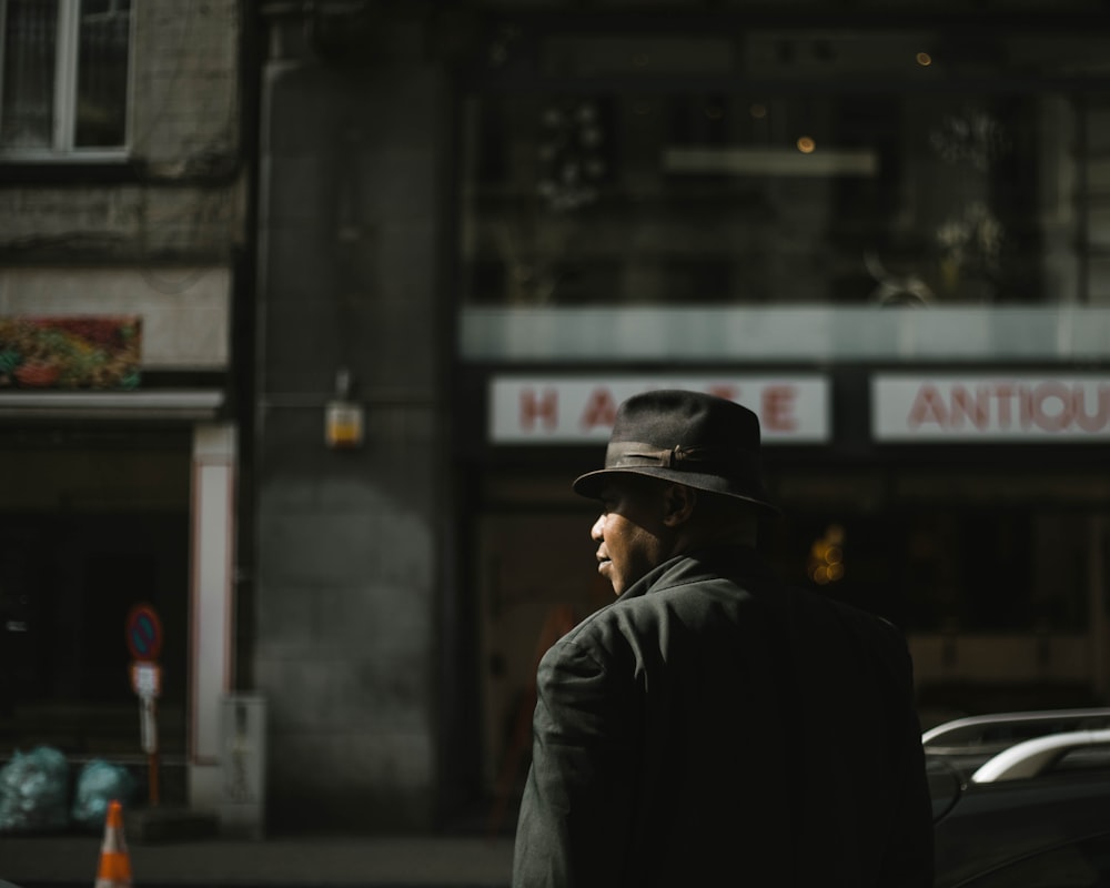 homem em jaqueta preta vestindo chapéu preto em pé perto do edifício durante o dia