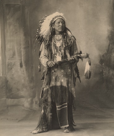 A sepia-toned portrait of a person dressed in traditional Native American attire. The individual is wearing a feathered headdress and a fringed garment adorned with beadwork and patterns. They are holding a decorated staff and standing in front of a blurred, indistinct background.