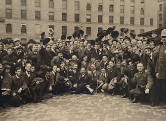 A large group of men dressed in early 20th century formal attire, including suits, coats, and bowler hats, gather closely together in an outdoor setting. The men display a variety of expressions, ranging from solemn to cheerful. Many hold their hats in the air. The background features the facade of a multi-story brick building with numerous windows, some of which are covered.