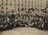 A large group of men dressed in early 20th century formal attire, including suits, coats, and bowler hats, gather closely together in an outdoor setting. The men display a variety of expressions, ranging from solemn to cheerful. Many hold their hats in the air. The background features the facade of a multi-story brick building with numerous windows, some of which are covered.
