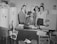 A group of four people are in an office setting, with three standing behind a desk where a man is sitting. The room contains various items including folders, documents, and promotional materials for products and services related to heating and utilities. Vintage appliances like water heaters and vacuum cleaners are visible, and the setting appears to be from the mid-20th century. The individuals are dressed in formal attire typical of the era.