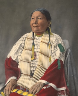 A person is adorned in traditional attire with intricate beadwork and distinctive patterns. The clothing features bright colors, including red and white with detailed decorations. The individual has long hair styled with accessories and appears poised and dignified.