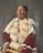 A person is adorned in traditional attire with intricate beadwork and distinctive patterns. The clothing features bright colors, including red and white with detailed decorations. The individual has long hair styled with accessories and appears poised and dignified.