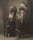Two individuals are wearing traditional Native American clothing, including feathered headdresses, standing side by side. One holds a rifle, while both have a calm and dignified expression. The image is sepia-toned, giving it a historical feel.