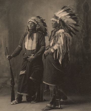 Two individuals are wearing traditional Native American clothing, including feathered headdresses, standing side by side. One holds a rifle, while both have a calm and dignified expression. The image is sepia-toned, giving it a historical feel.
