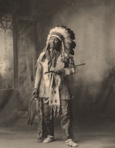 A person dressed in traditional indigenous attire stands indoors. The attire includes a feathered headdress, embroidered clothing, and patterned moccasins. The person is holding a long pipe and a fan made of feathers. The background features a painted backdrop with a window on the left side.