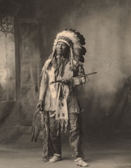 A person dressed in traditional indigenous attire stands indoors. The attire includes a feathered headdress, embroidered clothing, and patterned moccasins. The person is holding a long pipe and a fan made of feathers. The background features a painted backdrop with a window on the left side.