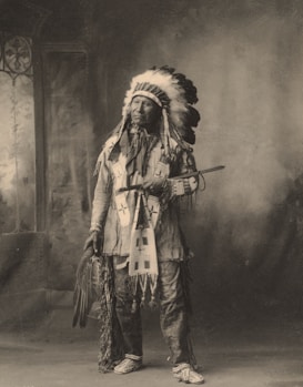 A person dressed in traditional indigenous attire stands indoors. The attire includes a feathered headdress, embroidered clothing, and patterned moccasins. The person is holding a long pipe and a fan made of feathers. The background features a painted backdrop with a window on the left side.