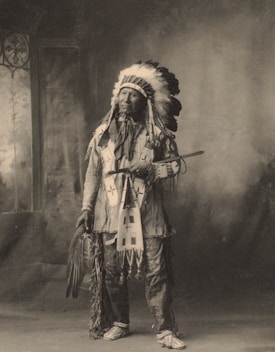 A person dressed in traditional indigenous attire stands indoors. The attire includes a feathered headdress, embroidered clothing, and patterned moccasins. The person is holding a long pipe and a fan made of feathers. The background features a painted backdrop with a window on the left side.