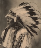 A sepia-toned portrait of a person wearing a traditional headdress with numerous feathers. The attire is adorned with intricate patterns and textures, highlighting cultural significance.