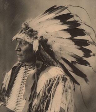 A sepia-toned portrait of a person wearing a traditional headdress with numerous feathers. The attire is adorned with intricate patterns and textures, highlighting cultural significance.