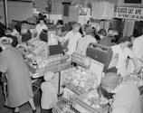 A busy grocery store with several people shopping and staff members assisting at the checkout counters. The area is filled with various products, including canned goods and cigarettes. Shoppers consist of both adults and children, with some employees dressed in white uniforms. Signs indicate the store will be closed for Patriot's Day.