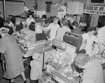 A busy grocery store with several people shopping and staff members assisting at the checkout counters. The area is filled with various products, including canned goods and cigarettes. Shoppers consist of both adults and children, with some employees dressed in white uniforms. Signs indicate the store will be closed for Patriot's Day.