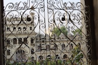 Elaborate wrought iron gates with intricate swirling designs frame a view of an elegant old building with arched windows and ornate architectural features. Greenery is visible through the openings, adding a touch of natural beauty to the scene.