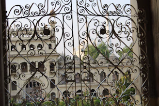 Elaborate wrought iron gates with intricate swirling designs frame a view of an elegant old building with arched windows and ornate architectural features. Greenery is visible through the openings, adding a touch of natural beauty to the scene.