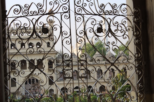 Elaborate wrought iron gates with intricate swirling designs frame a view of an elegant old building with arched windows and ornate architectural features. Greenery is visible through the openings, adding a touch of natural beauty to the scene.