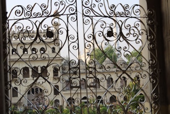 Elaborate wrought iron gates with intricate swirling designs frame a view of an elegant old building with arched windows and ornate architectural features. Greenery is visible through the openings, adding a touch of natural beauty to the scene.