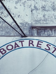 A weathered wall with peeling paint and visible cracks in a rustic setting. The letters 'BOAT REST' are partially visible, painted in dark red and surrounded by a faded blue arc. Electrical wires and metal rods are attached to the wall.