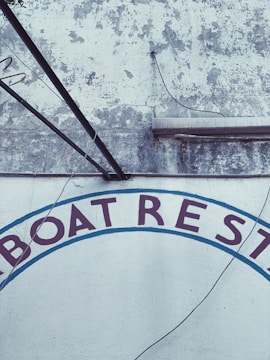 A weathered wall with peeling paint and visible cracks in a rustic setting. The letters 'BOAT REST' are partially visible, painted in dark red and surrounded by a faded blue arc. Electrical wires and metal rods are attached to the wall.