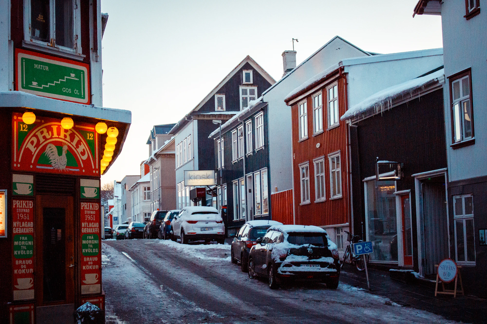 A brightly lit bar storefront opens onto a snowy Reykjavik street lined with colorful buildings.