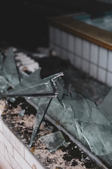 Several large and jagged pieces of broken glass are scattered on a tiled surface, with some pieces leaning against each other. The surface shows signs of wear and weathering, suggesting an abandoned or neglected area.
