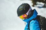 A person wearing a black helmet and reflective ski goggles stands outdoors. The individual is dressed in a bright blue ski jacket and appears to be engaged in a winter sport activity on a snowy terrain.