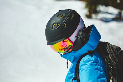A person wearing a black helmet and reflective ski goggles stands outdoors. The individual is dressed in a bright blue ski jacket and appears to be engaged in a winter sport activity on a snowy terrain.