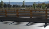 A road with a concrete barrier and yellow railings in the foreground, leading to an area with trees and a fence. In the distance, mountains can be seen under a clear blue sky. Shadows from the railings fall onto the pavement, and a vehicle is parked near the fence.