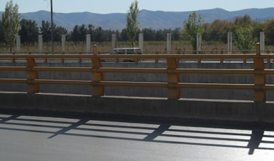 A road with a concrete barrier and yellow railings in the foreground, leading to an area with trees and a fence. In the distance, mountains can be seen under a clear blue sky. Shadows from the railings fall onto the pavement, and a vehicle is parked near the fence.