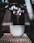 A close-up shot of a coffee machine espresso spout pouring a shot of coffee into a white cup with a logo. The setup includes metallic textures of the machine and soft focus highlights in the background.