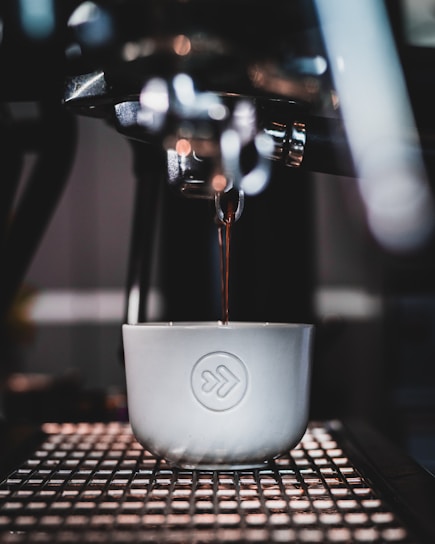 A close-up shot of a coffee machine espresso spout pouring a shot of coffee into a white cup with a logo. The setup includes metallic textures of the machine and soft focus highlights in the background.
