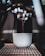A close-up shot of a coffee machine espresso spout pouring a shot of coffee into a white cup with a logo. The setup includes metallic textures of the machine and soft focus highlights in the background.