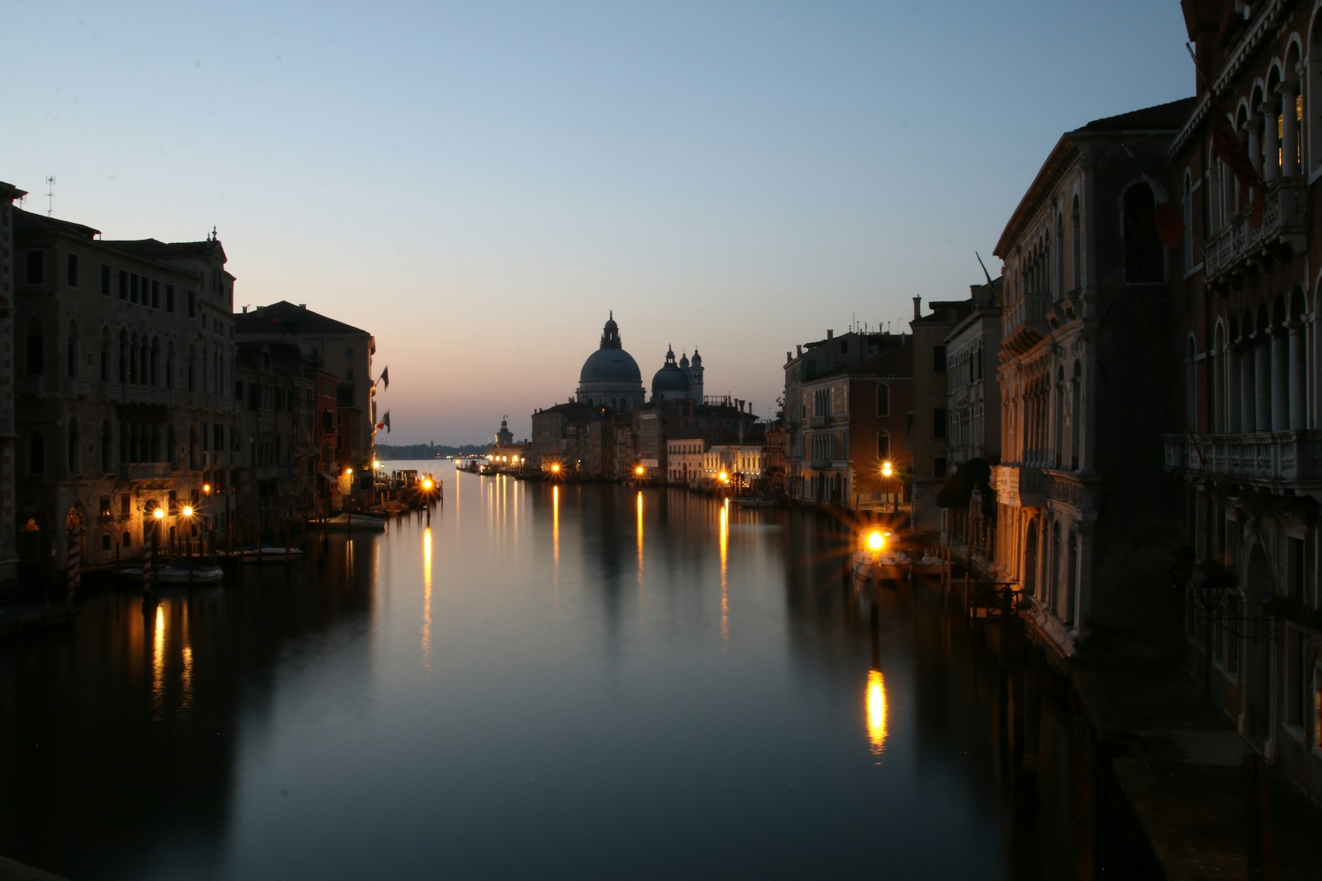 A serene evening view of the Navigli canals at sunset, with reflections of colorful buildings and softly glowing street lamps.