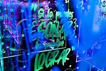 Bright neon lights create an energetic and lively atmosphere with glowing text and decorative symbols filling the space. The text, in Spanish, conveys an inspirational message amidst a backdrop of stars and abstract patterns.